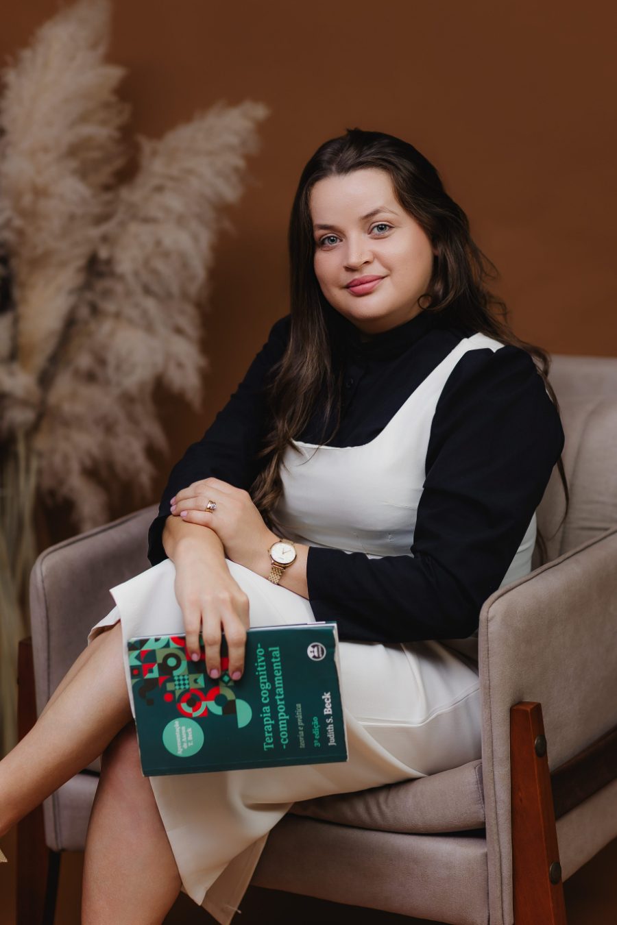 Claudiane Albarracin, psicóloga, sentada em uma poltrona com um livro de Terapia Cognitivo-Comportamental nas mãos.